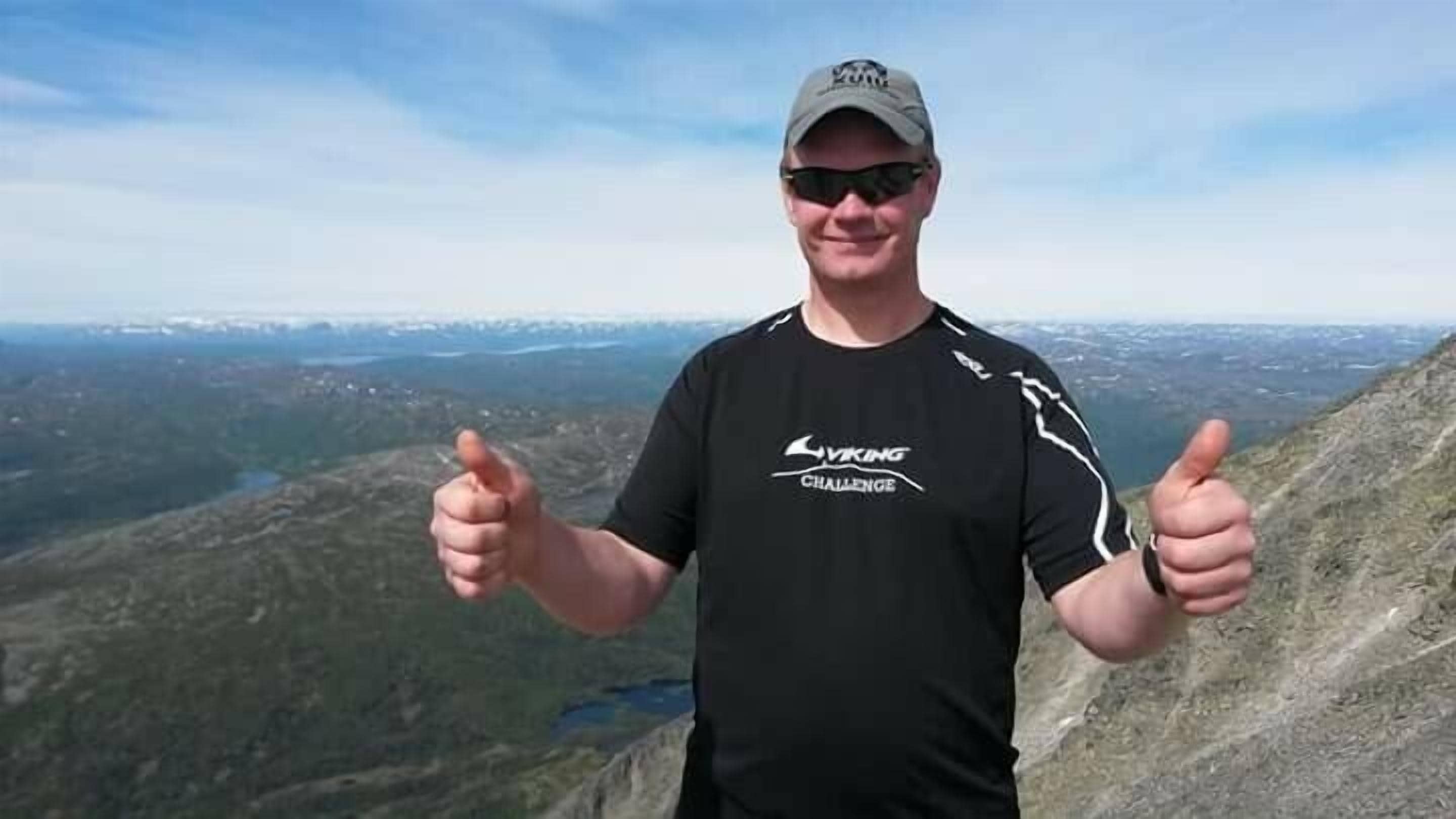 A person wearing a black t-shirt with a logo and text, standing on a mountain with a scenic view in the background.