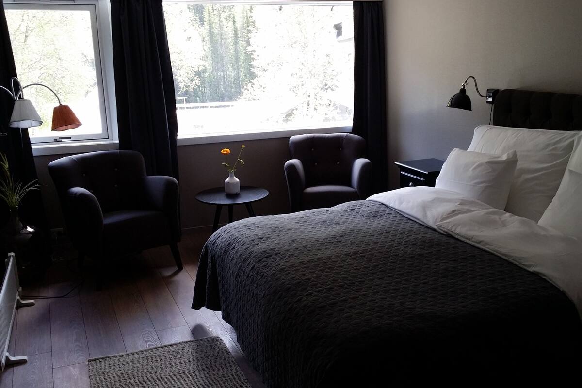 Een hotelkamer met een bed, twee stoelen, een kleine tafel met een vaas en een raam met gordijnen.