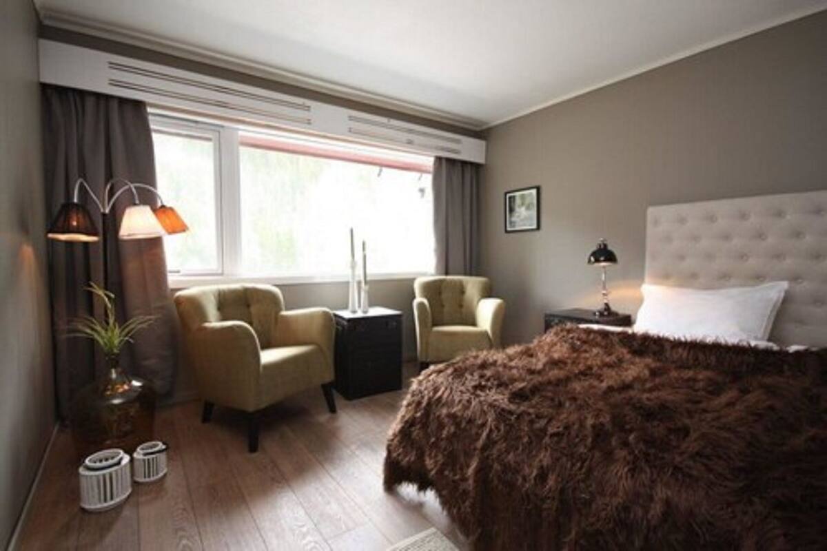 Een slaapkamer met een groot bed, twee fauteuils, een bijzettafeltje, een lamp en een raam met gordijnen.