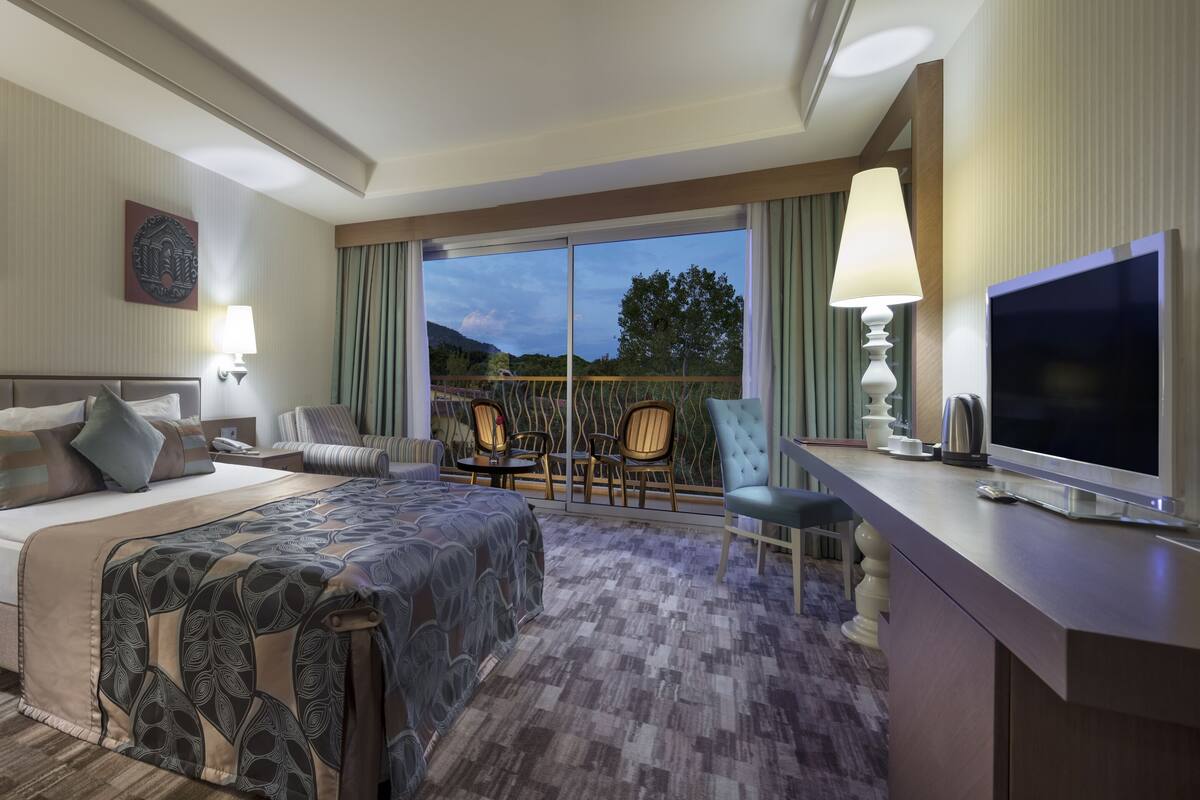A hotel room with a large bed, a desk with a TV, a balcony with chairs, and artwork on the walls.