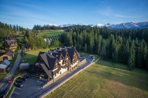 Una cabaña con estilo arquitectónico alpino tradicional, con un edificio central de techo inclinado y múltiples ventanas. Hay un área de estacionamiento con varios autos estacionados al frente, y el paisaje circundante incluye una zona boscosa con vista a montañas distantes.
