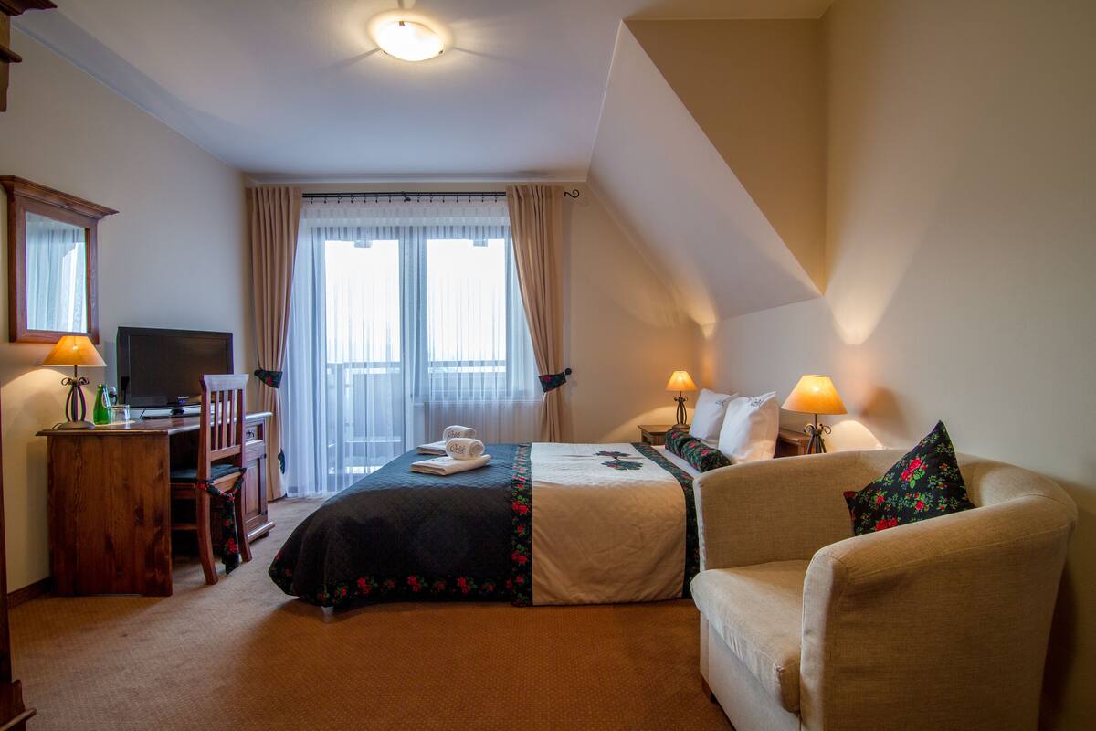 Habitación de hotel con una cama, dos mesitas de noche, una silla, un escritorio con una computadora y una televisión.