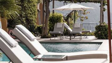 Zona junto a la piscina con sillones de descanso y sombrillas, rodeada de follaje y un pérgola.