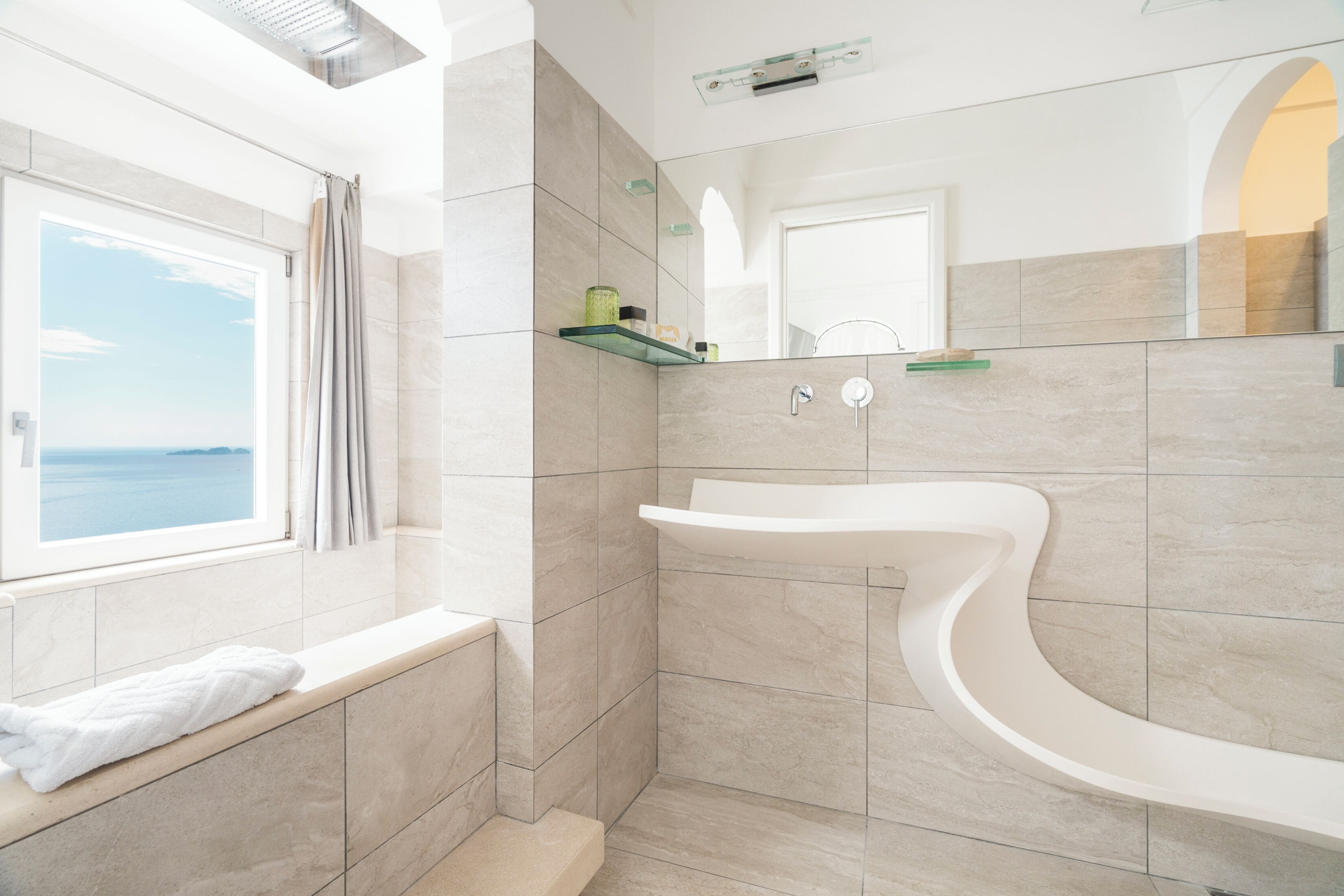 A modern bathroom with a curved sink, a window with a view of the sea, and a towel on the bench.