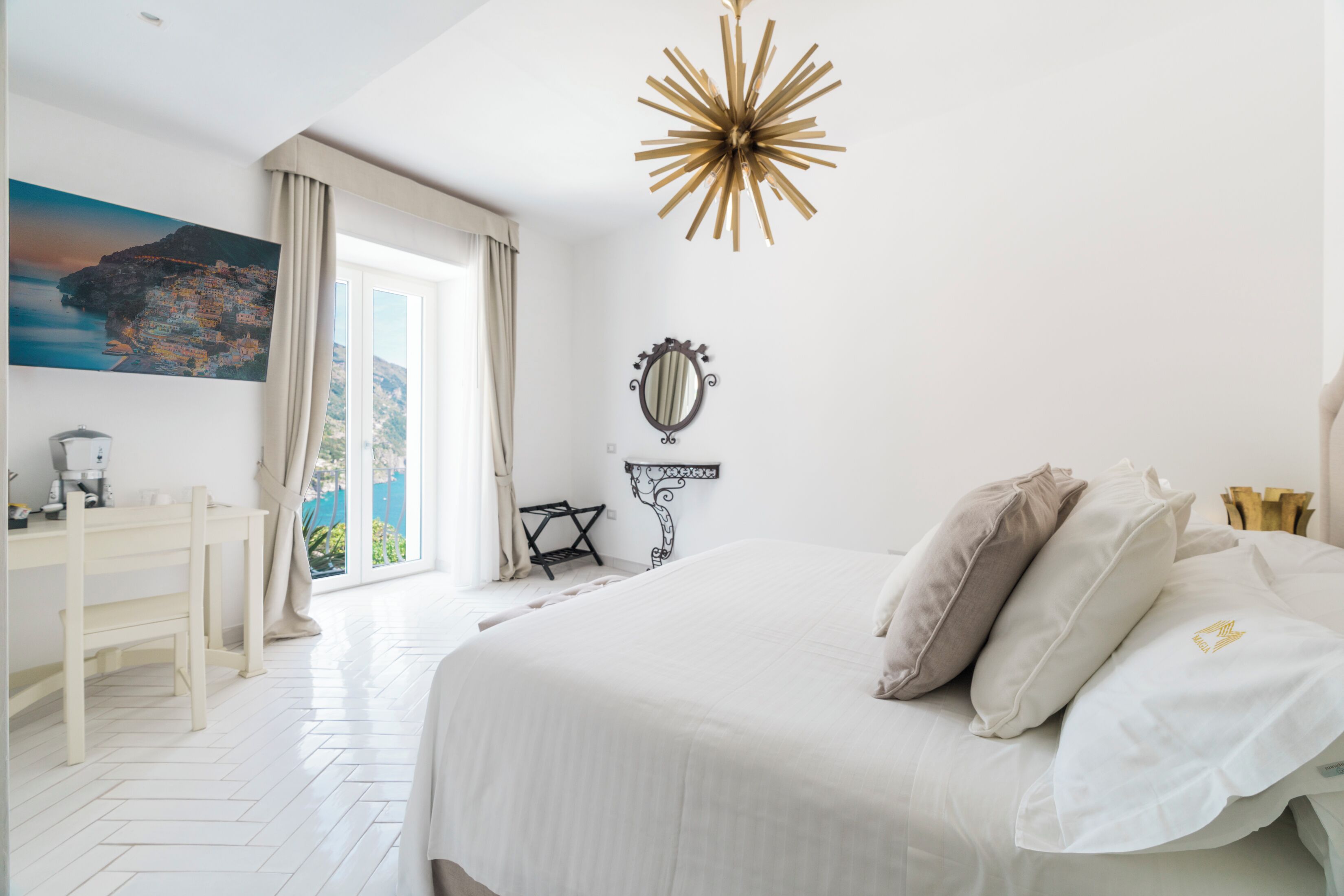 A bedroom with a large bed, a desk, a chair, a mirror, and a view of a coastal town through the window.