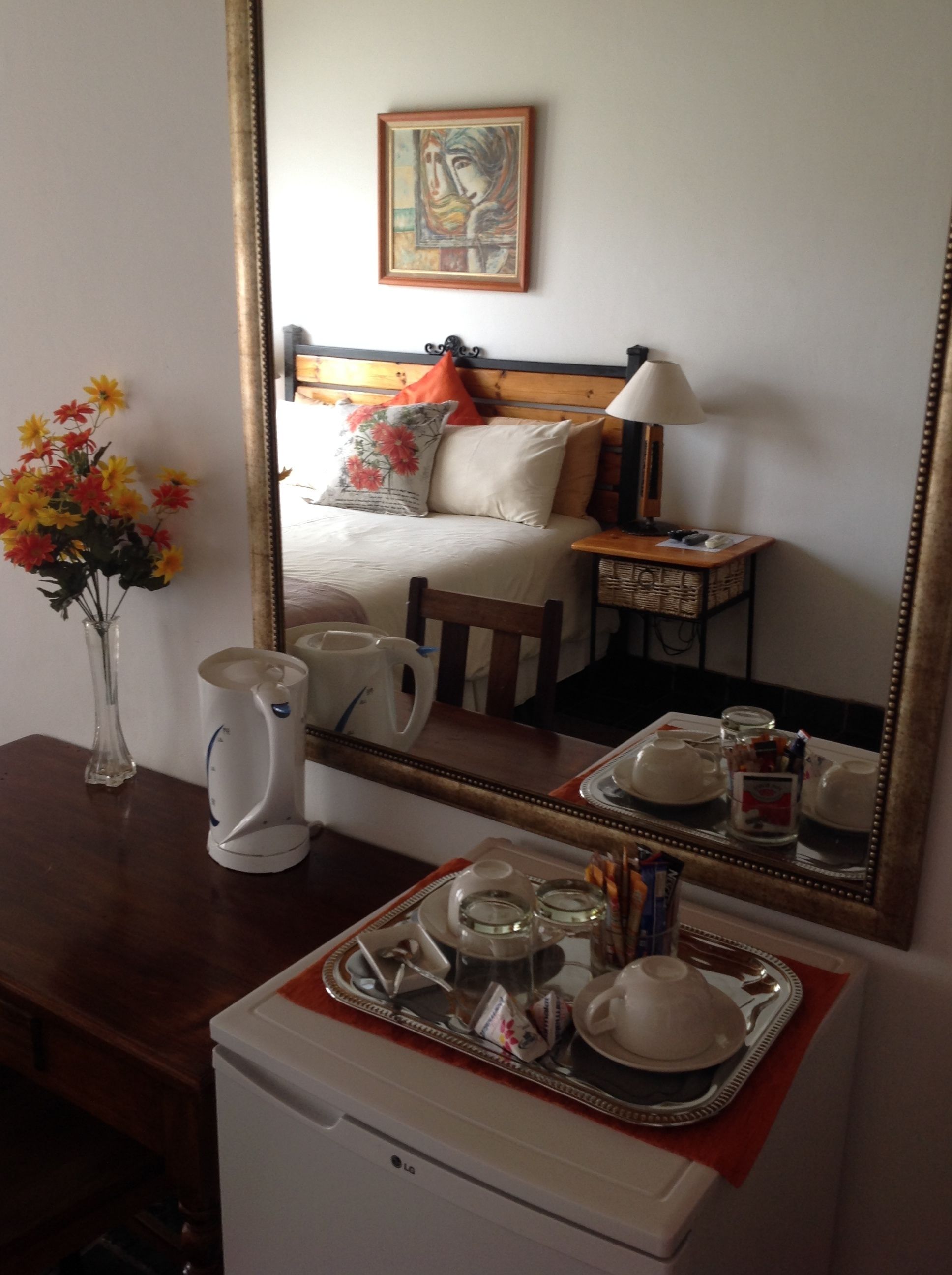 Quarto de hotel com cama, mesa de cabeceira, espelho, um vaso com flores, chaleira e bandeja com xícaras e pires.