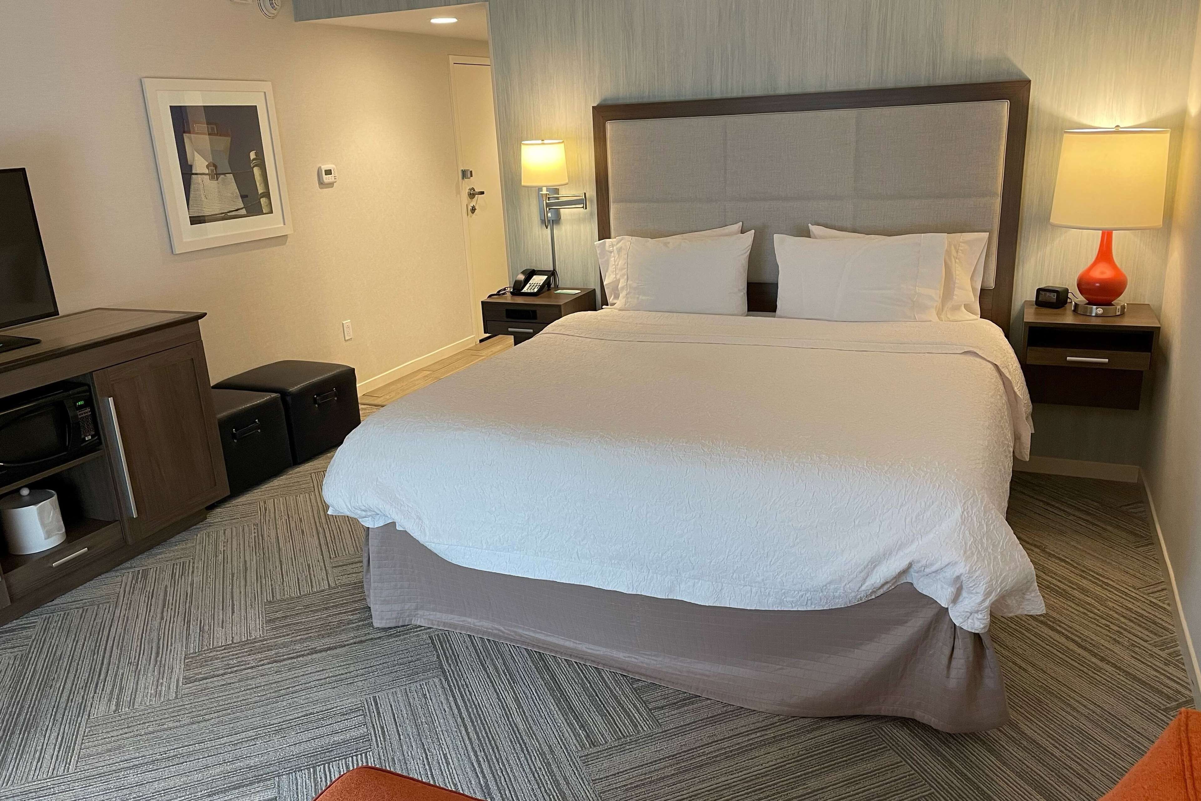 A hotel room with a large bed, a nightstand with a lamp, a desk with a computer, and a TV.