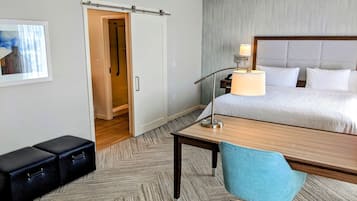 A hotel room with a bed, a desk, a chair, a nightstand, a lamp, a wardrobe, and a painting on the wall.