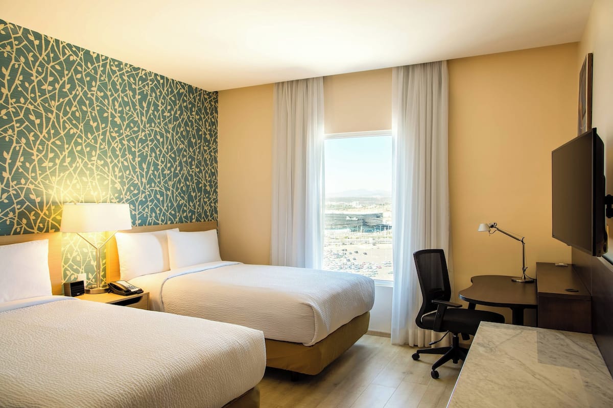A hotel room with two beds, a desk, a chair, a TV, and a window with a view.
