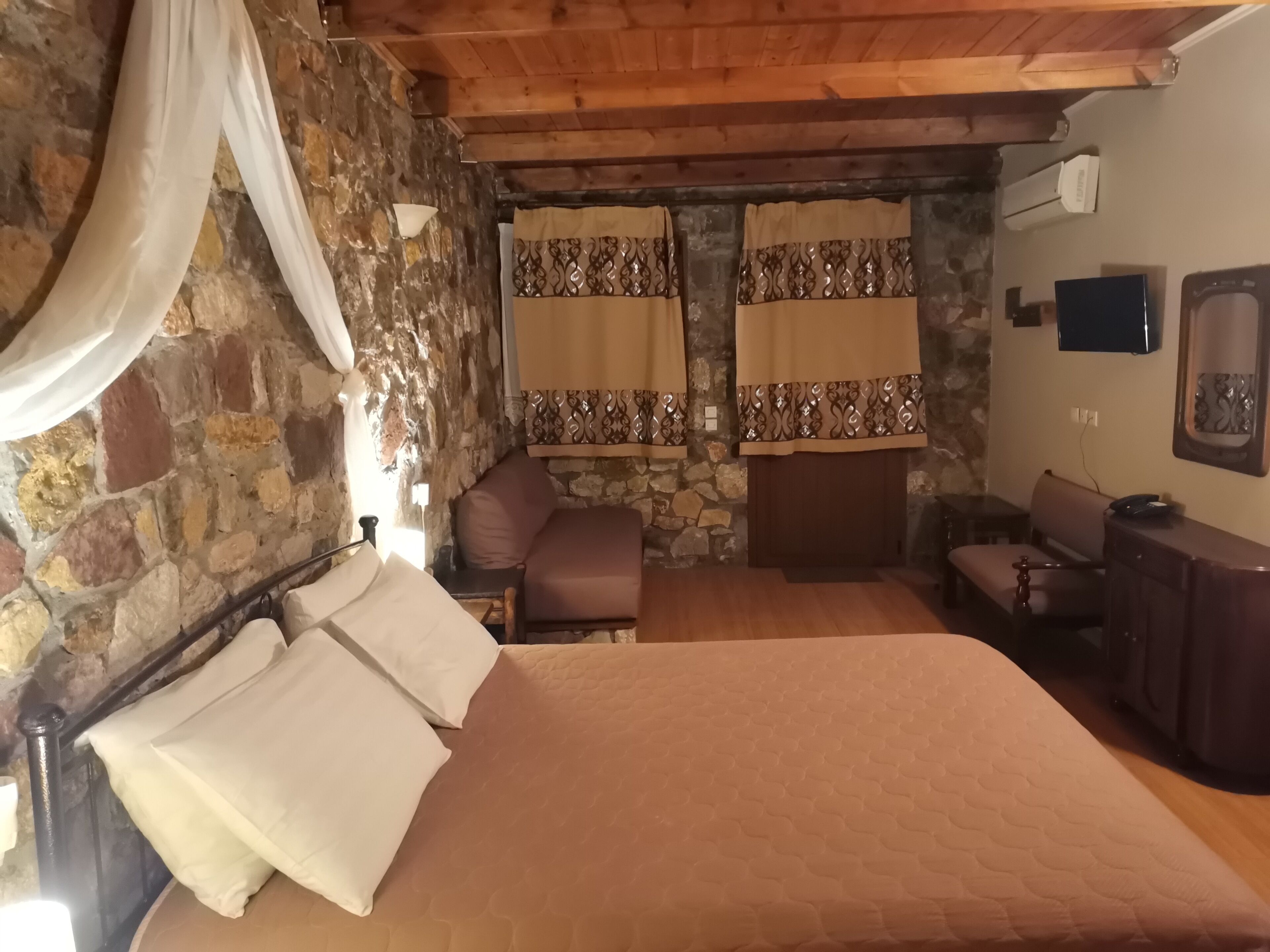 A bedroom with a stone wall, a bed with white and beige bedding, a wooden bench, and a television on the wall.