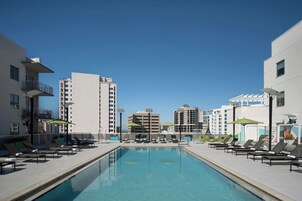 Piscina con sillones de descanso y sombrillas, rodeada de edificios modernos.