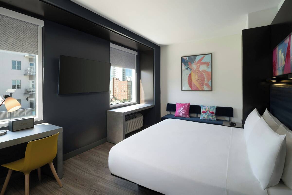 A modern hotel room with a bed, desk, chair, and artwork on the wall.