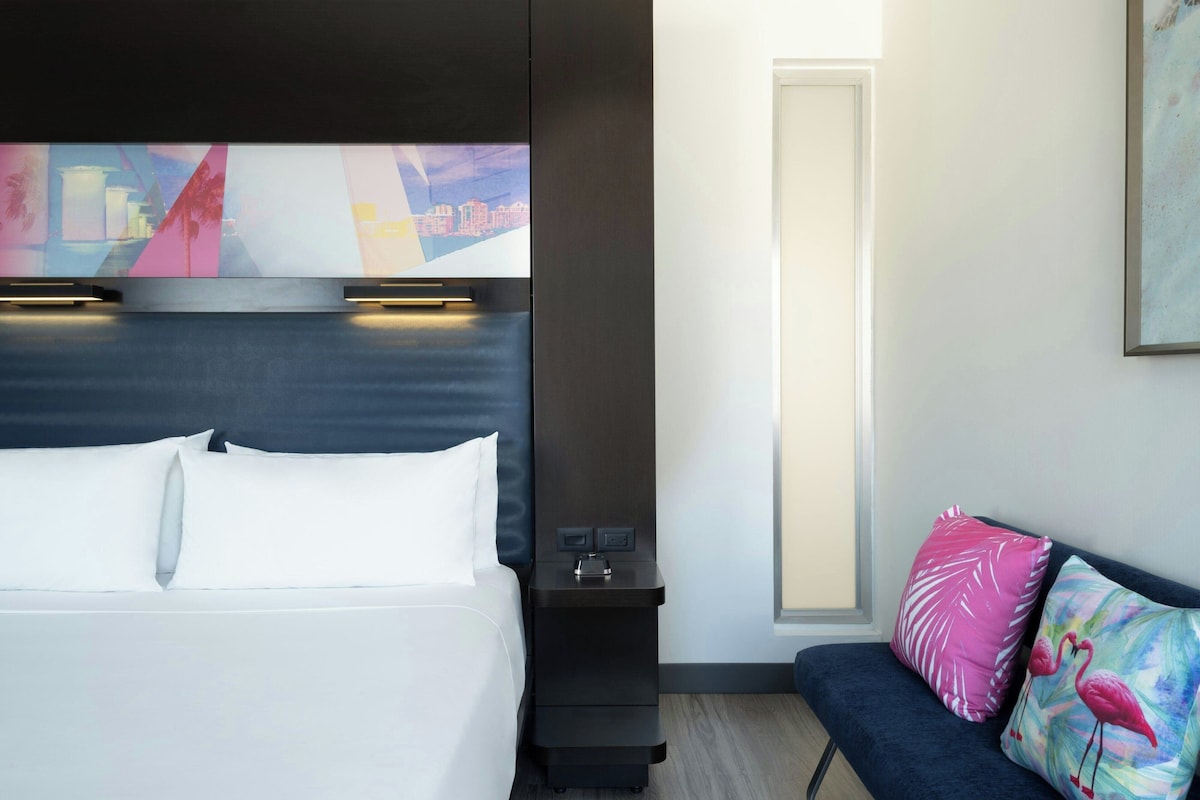 A hotel room with a bed, bedside table, nightstand, and a sofa with decorative pillows.
