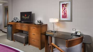 A hotel room with a wooden desk, a television, a minibar, a lamp, and a framed picture on the wall.