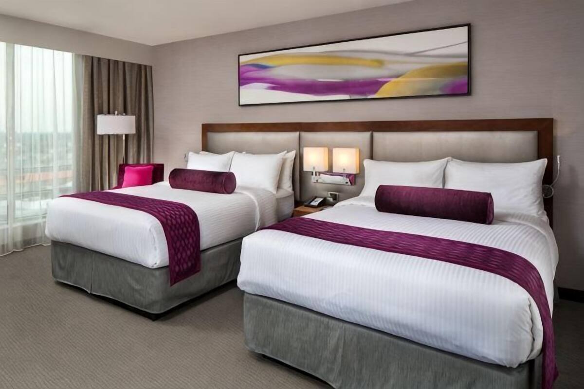 A hotel room with two beds, a large window, a lamp, and a painting on the wall.