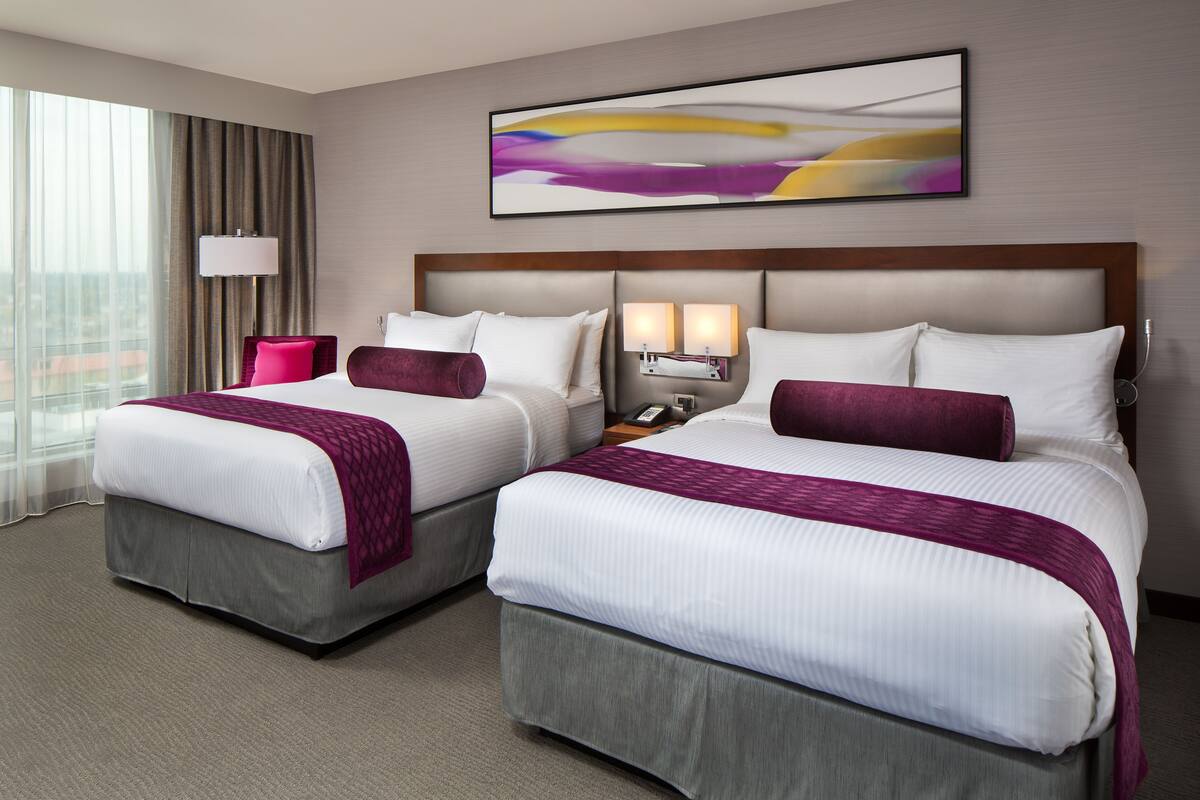 A hotel room with two beds, a large window with curtains, a wall-mounted abstract painting, and a nightstand with a lamp.