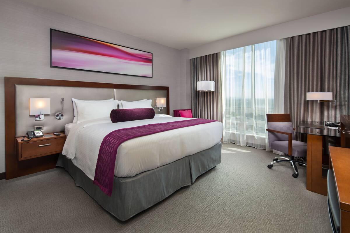 A hotel room with a large bed, a desk, a chair, and a view of the city through the window.