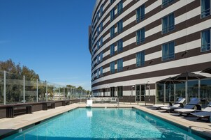 Un hotel moderno con piscina, tumbonas y un balcón con cristales.