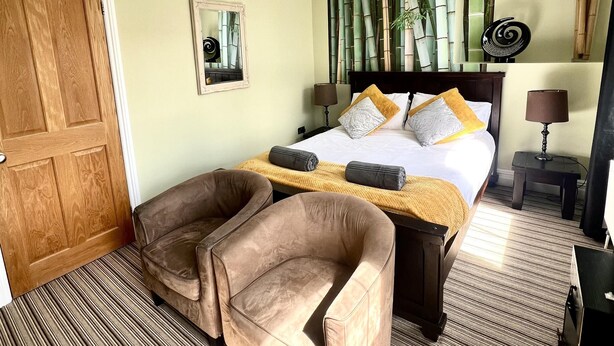 Habitación de hotel con cama, dos sillones, una puerta de madera, un espejo y una pared con estampado de bambú.