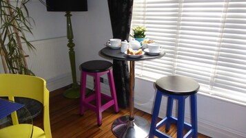 A room with a small round table set with a teapot, cups, and a plate of pastries. There are two stools, one pink and one blue, and a yellow chair. A potted plant is visible in the corner.