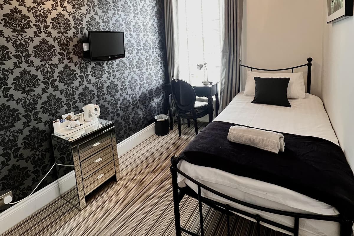 Ett hotellrum med en säng, ett nattduksbord med en telefon, ett skrivbord med en stol, ett fönster med gardiner och en väggmonterad TV.