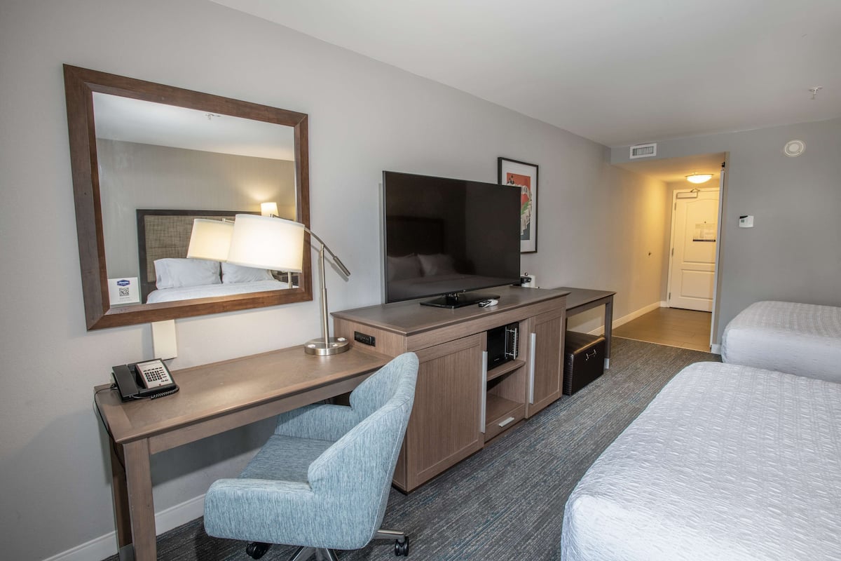 Ett hotellrum med ett skrivbord, en stol, en säng, en tv och en stor spegel.