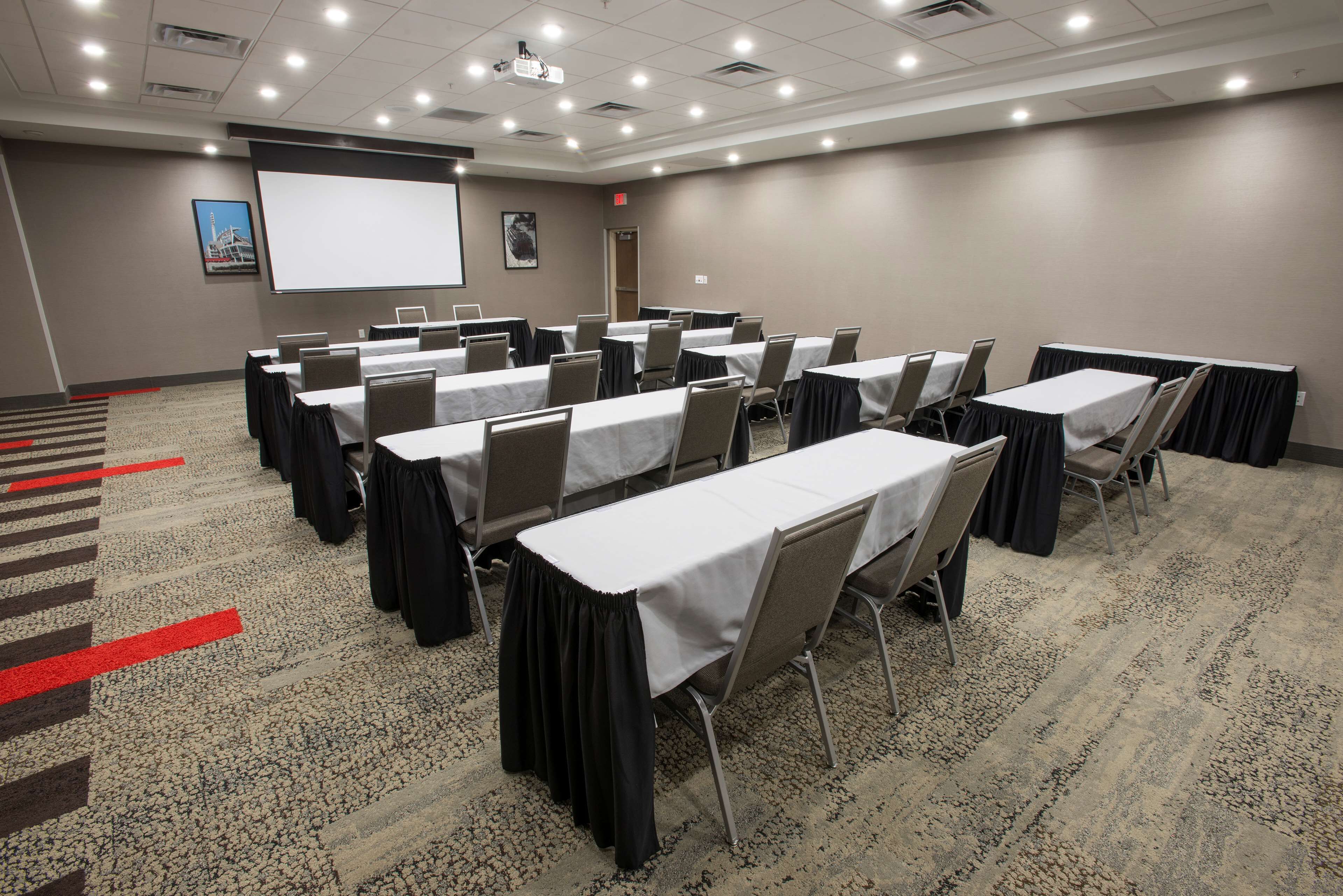 Una sala de conferencias con varias mesas y sillas, una pantalla de proyector y suelo alfombrado.