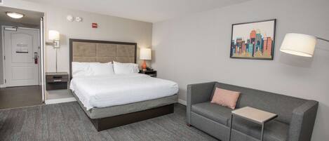 A hotel room with a bed, a gray sofa, a small table, a lamp, and a cityscape painting on the wall.