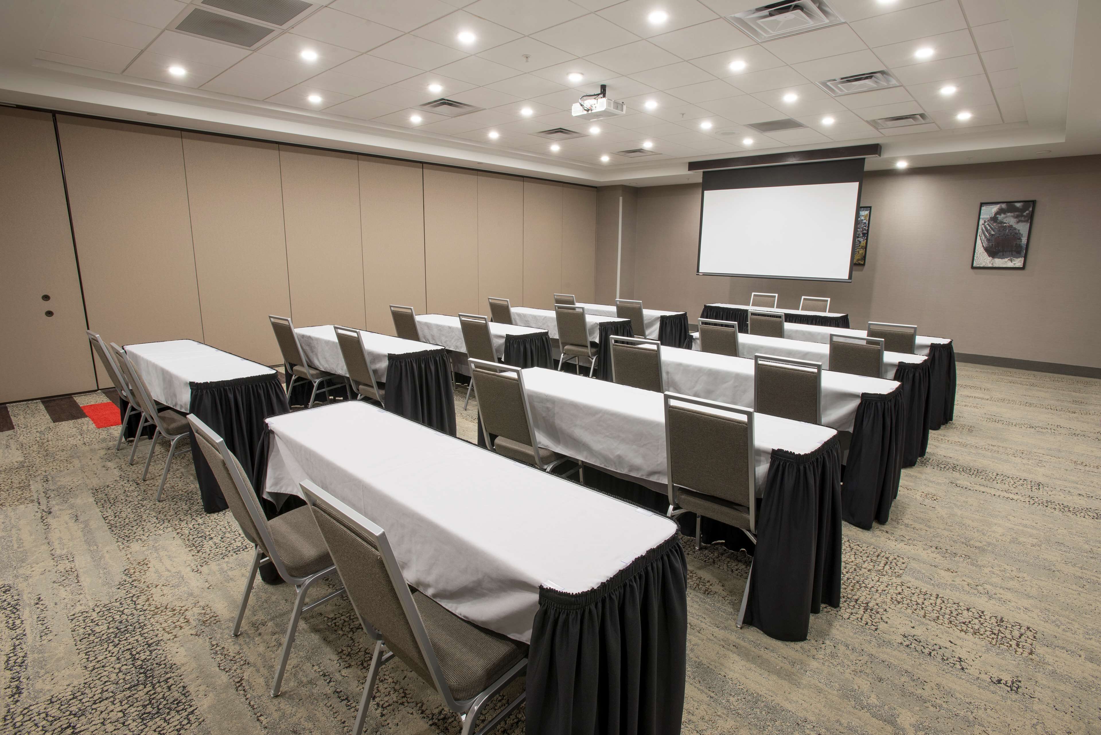 A conference room with multiple tables, chairs, a projector screen, and a carpeted floor.