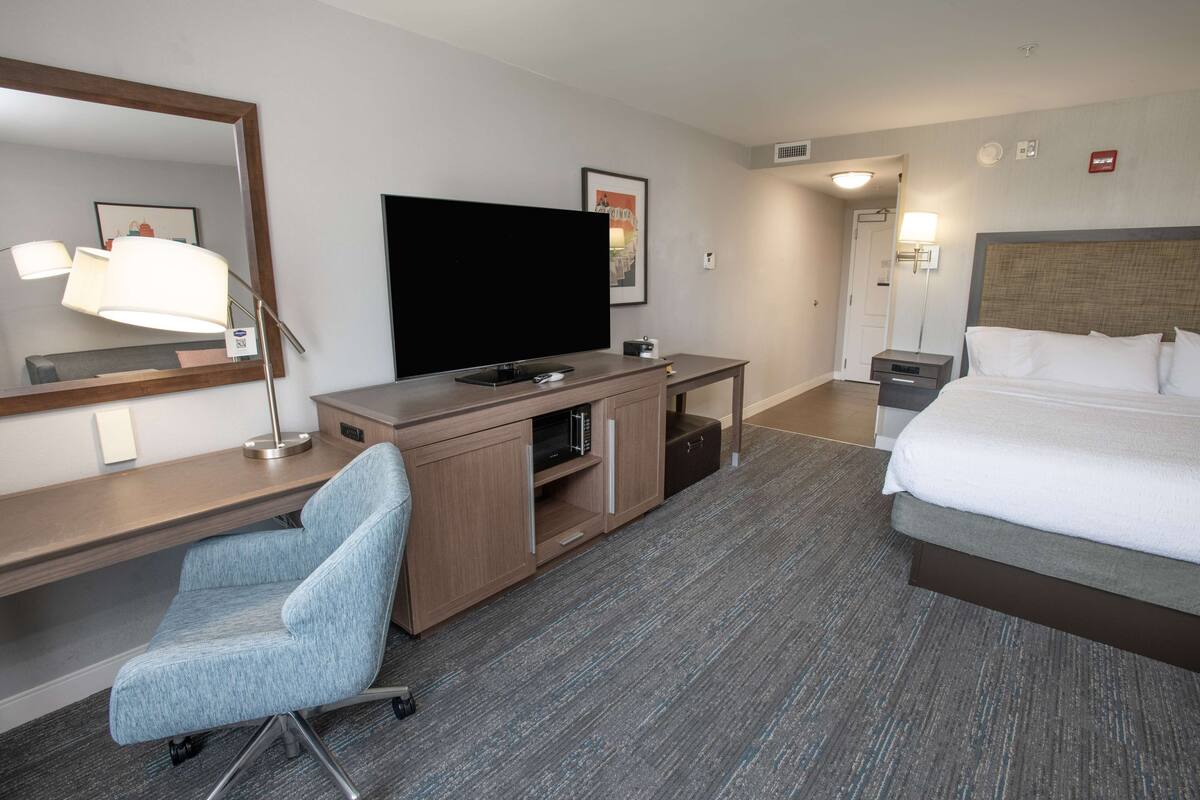 Ett hotellrum med en säng, ett skrivbord med en platt-TV, en stol, en spegel och en lampa.