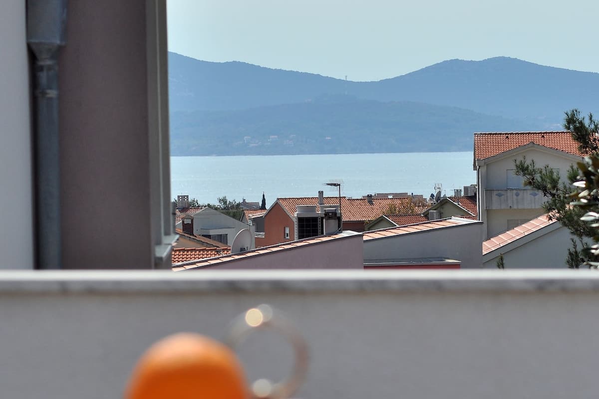 A table with a fruit bowl containing apples and oranges, and a view of a coastal town with buildings and mountains in the background.