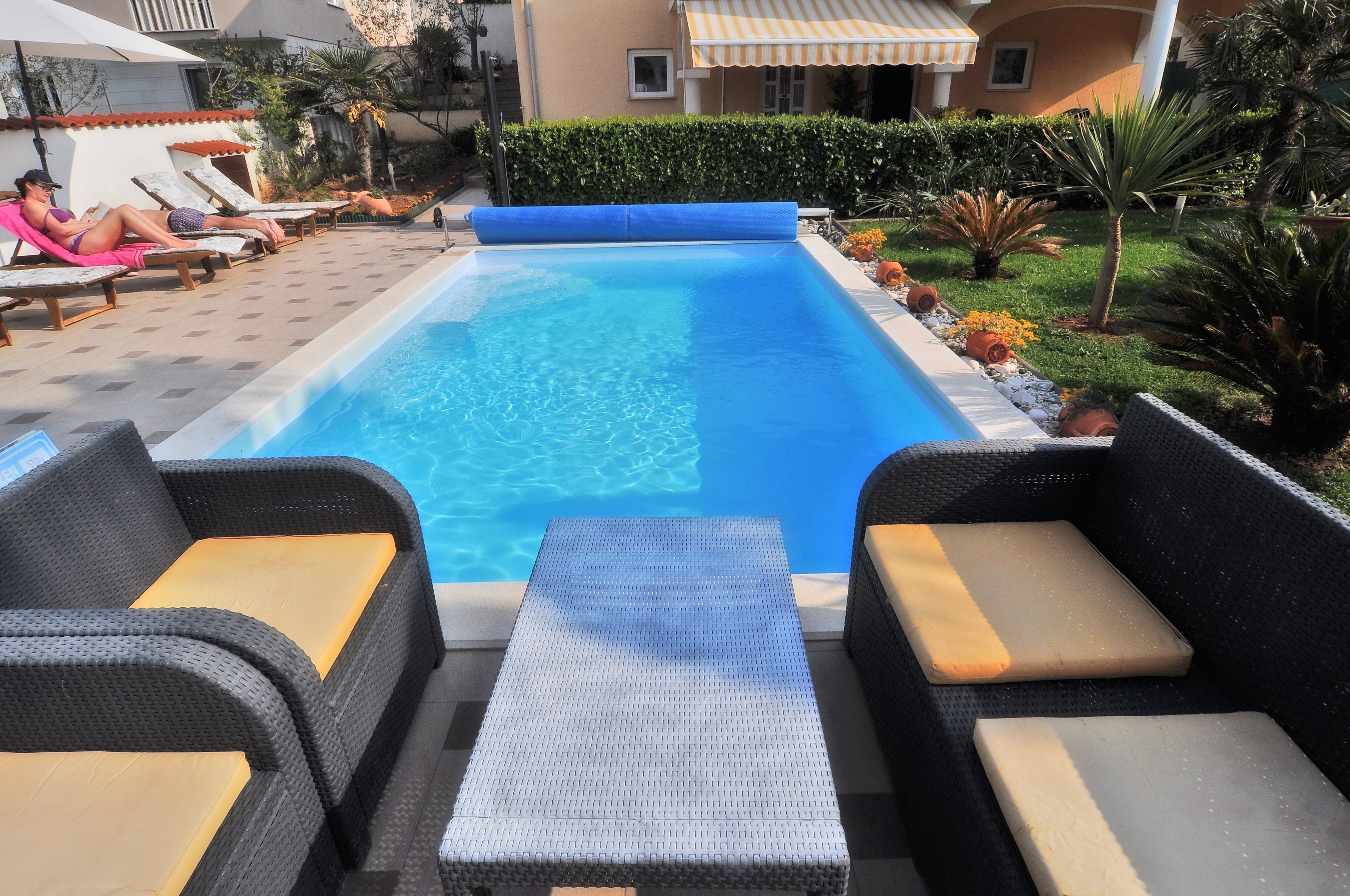 A swimming pool with lounge chairs and a table, surrounded by greenery and a building with a yellow awning.