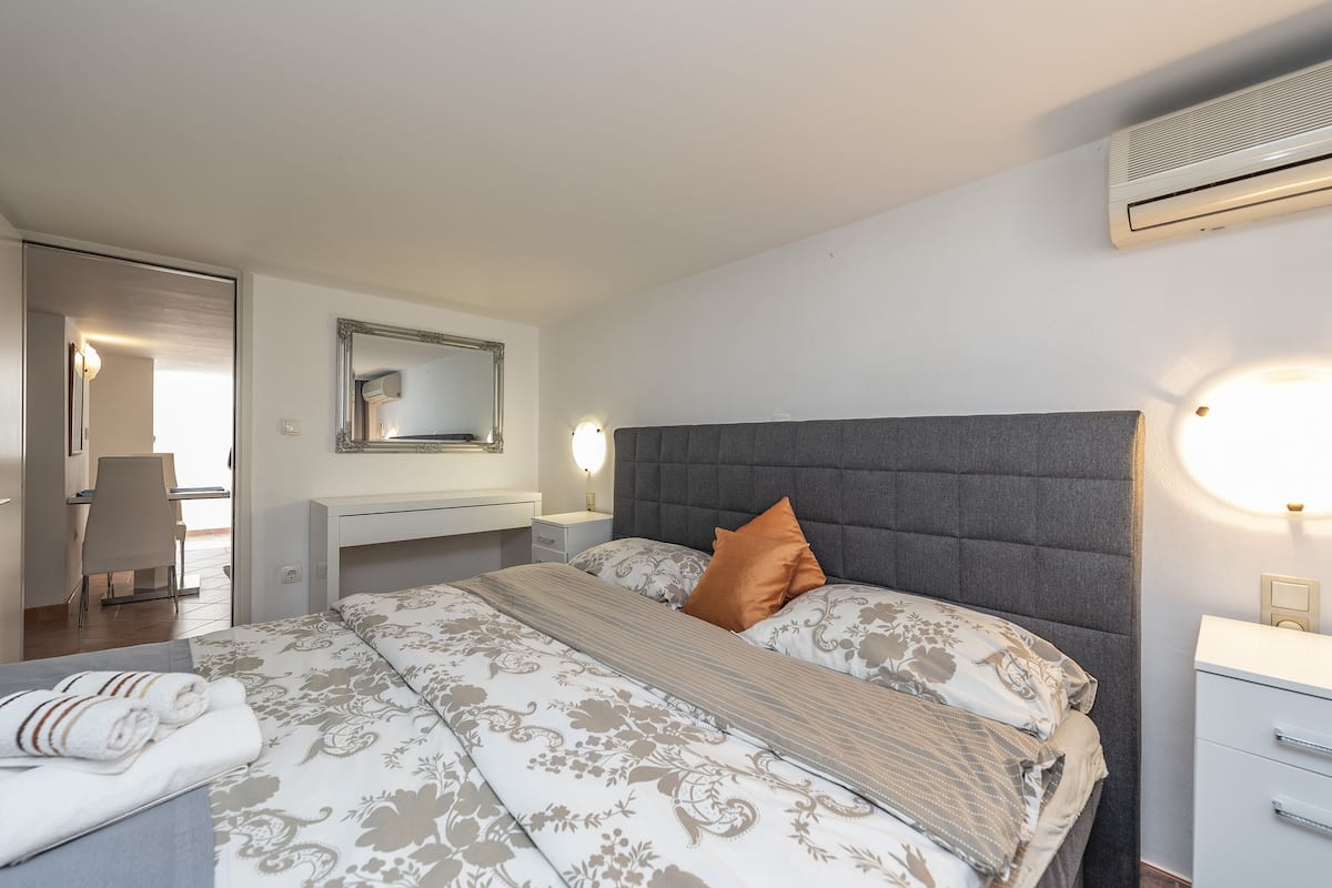 A neatly arranged bedroom with a bed, bedside tables, a mirror, and an air conditioning unit.