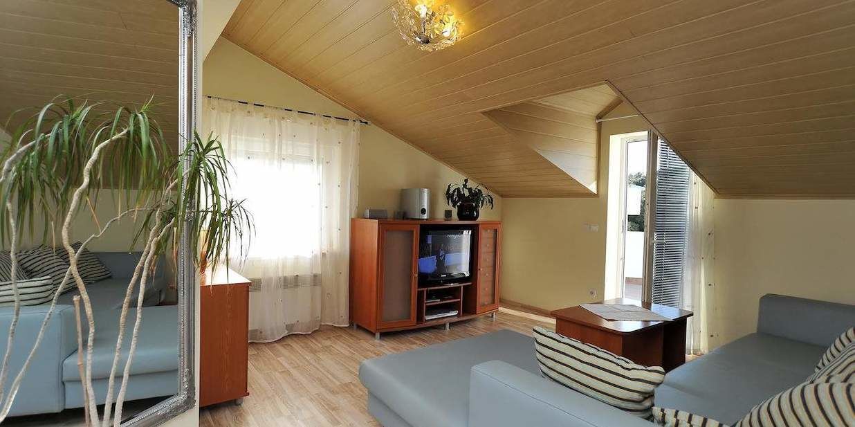 A cozy attic room with a wooden ceiling, a television, a sofa, and a large window.