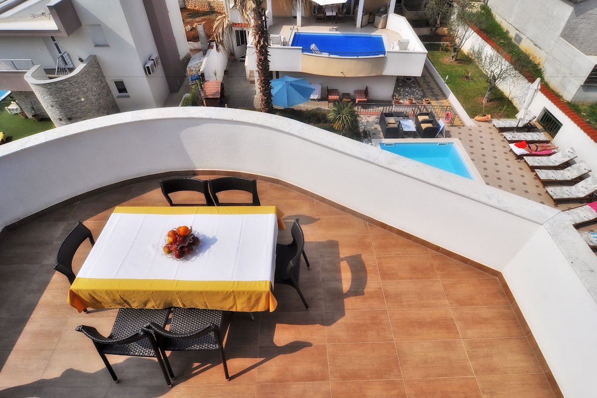 A rooftop terrace with a table set for two, surrounded by chairs, overlooking a swimming pool and a garden.