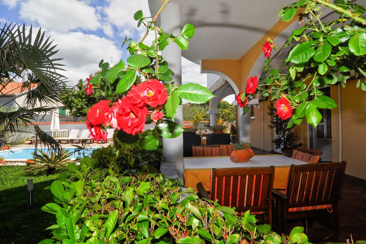 A poolside area with a covered seating area, a dining table, and lush greenery.