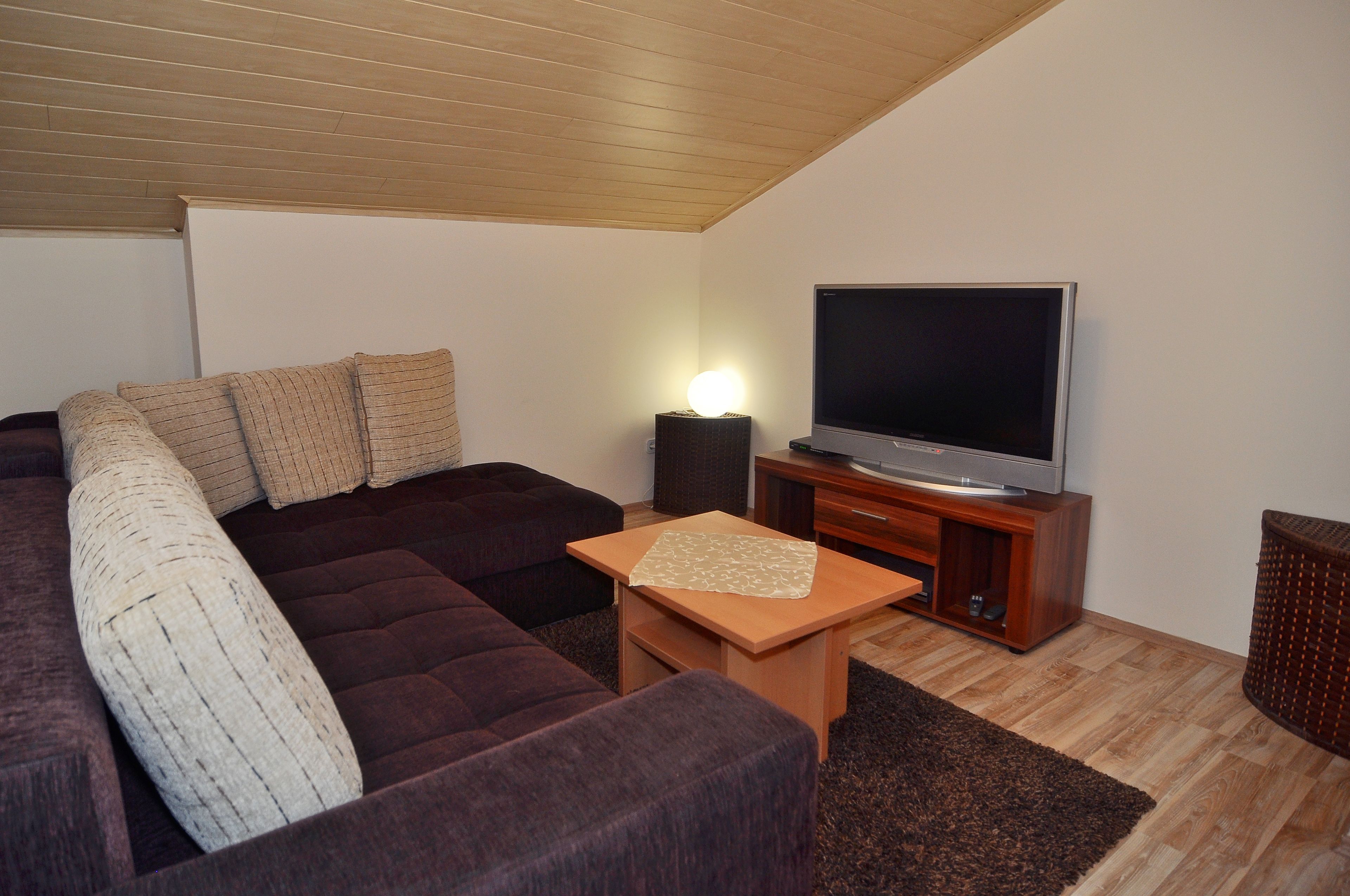 Una sala de estar con un sofá modular, un televisor de pantalla plana sobre un soporte de madera, una pequeña mesa de madera y una alfombra marrón oscuro.
