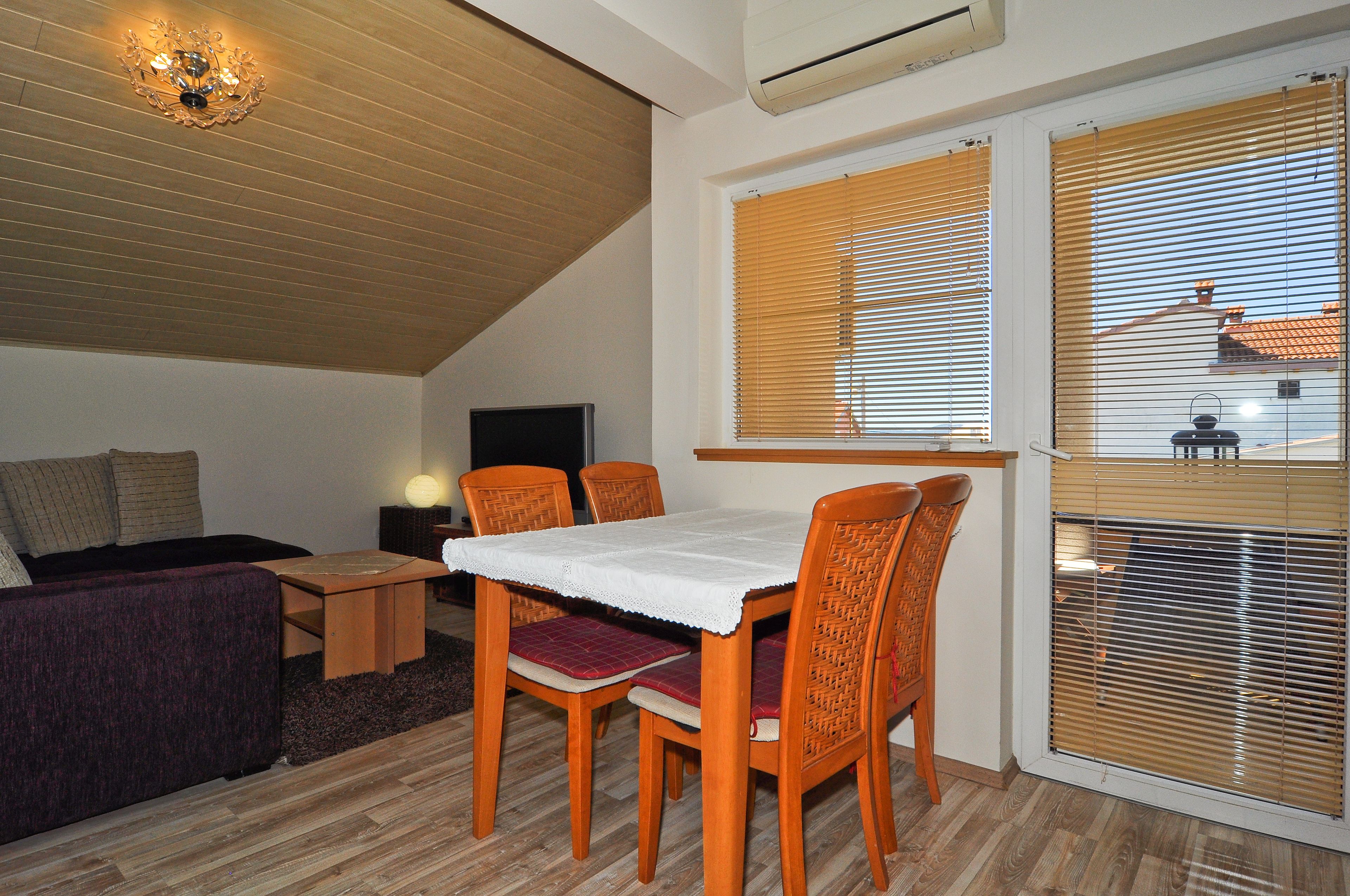 A cozy living room with a dining area, wooden ceiling, and large windows with blinds.