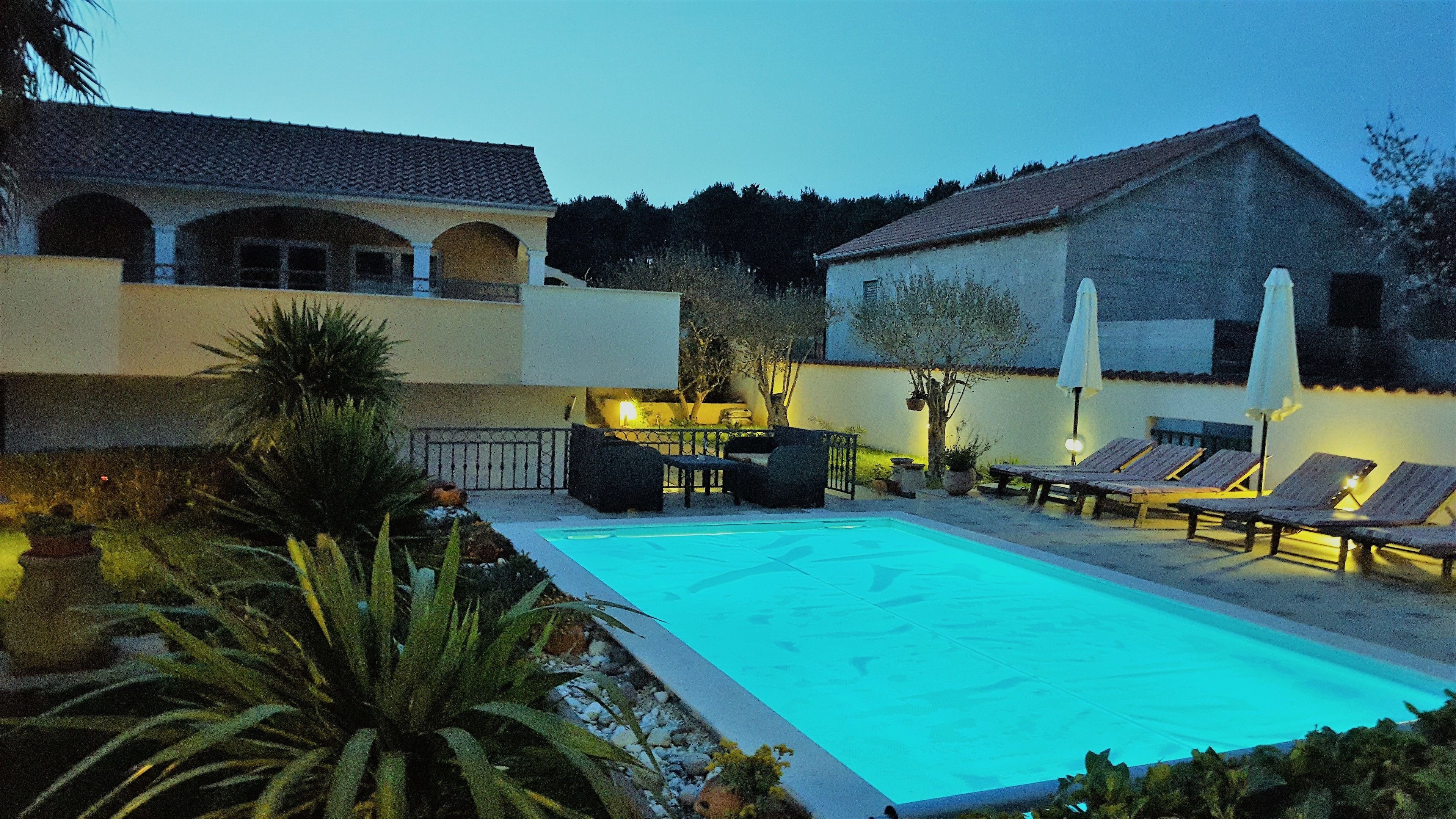 Une piscine entourée d’un jardin et d’un bâtiment à toit de tuiles, avec des chaises longues et une table à proximité.