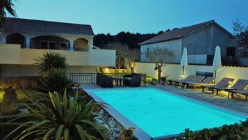 Une piscine entourée d’un jardin et d’un bâtiment à toit de tuiles, avec des chaises longues et une table à proximité.