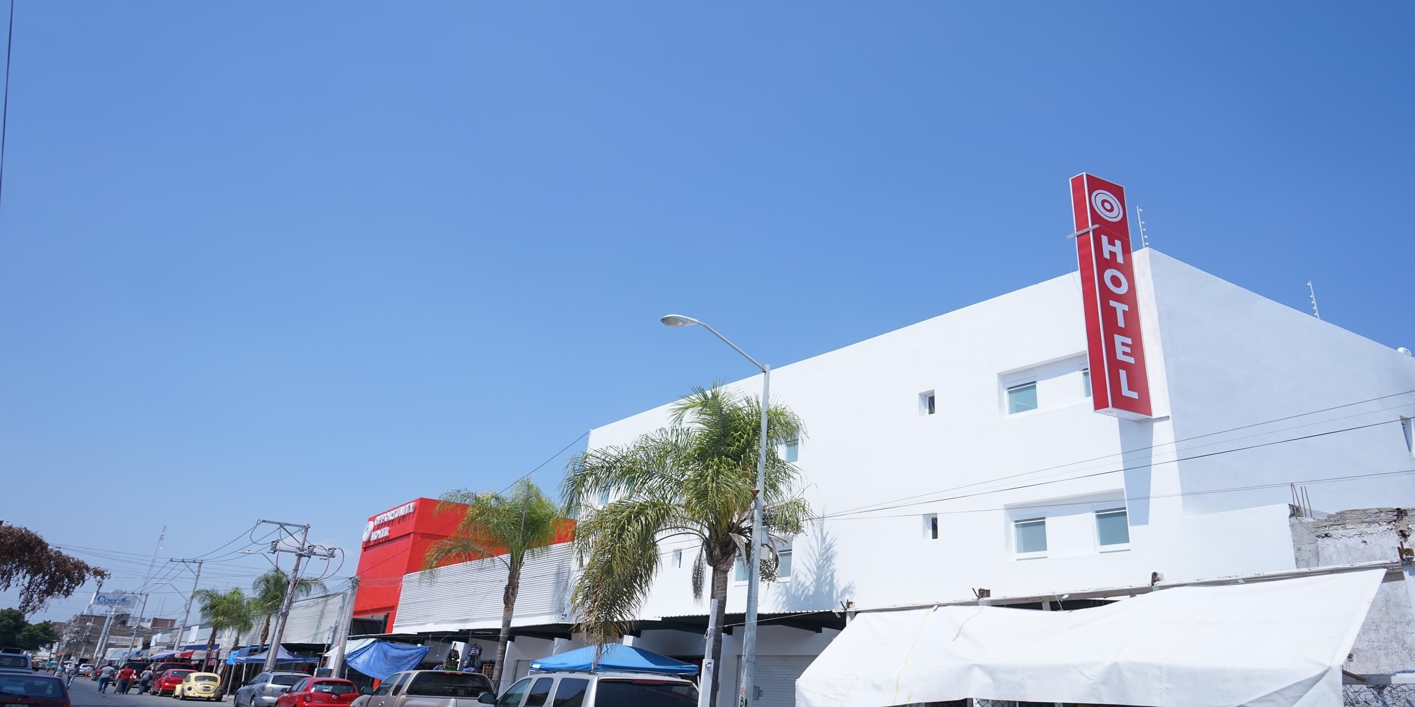 A white building with a red 'HOTEL' sign, a palm tree, and parked cars on the street.