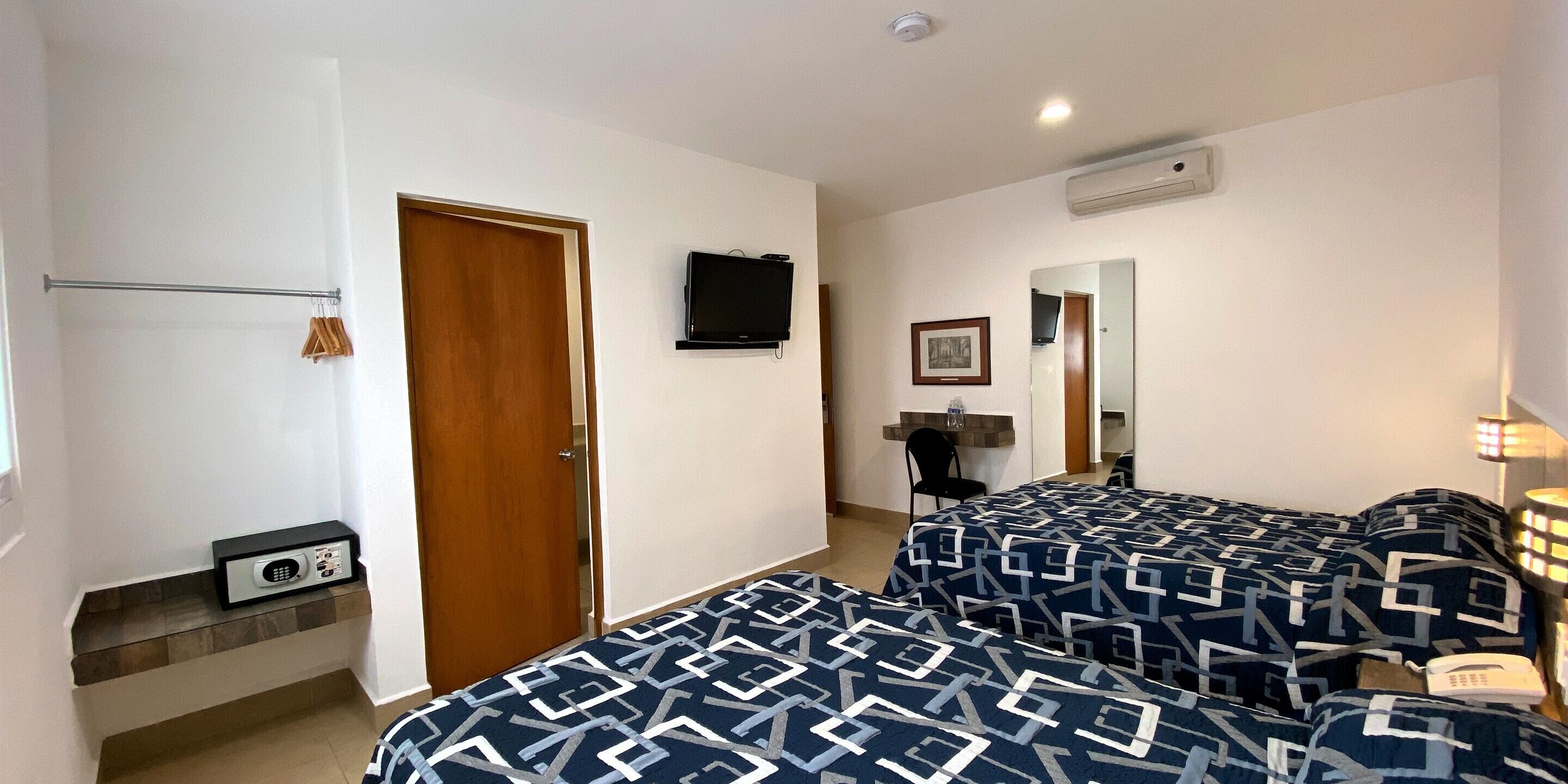 A hotel room with two beds, a TV, a desk with a chair, a mirror, and a wall-mounted heater.