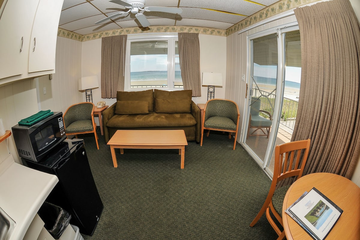 A compact room with a sofa, two chairs, a small table, a microwave, and a trash bin.