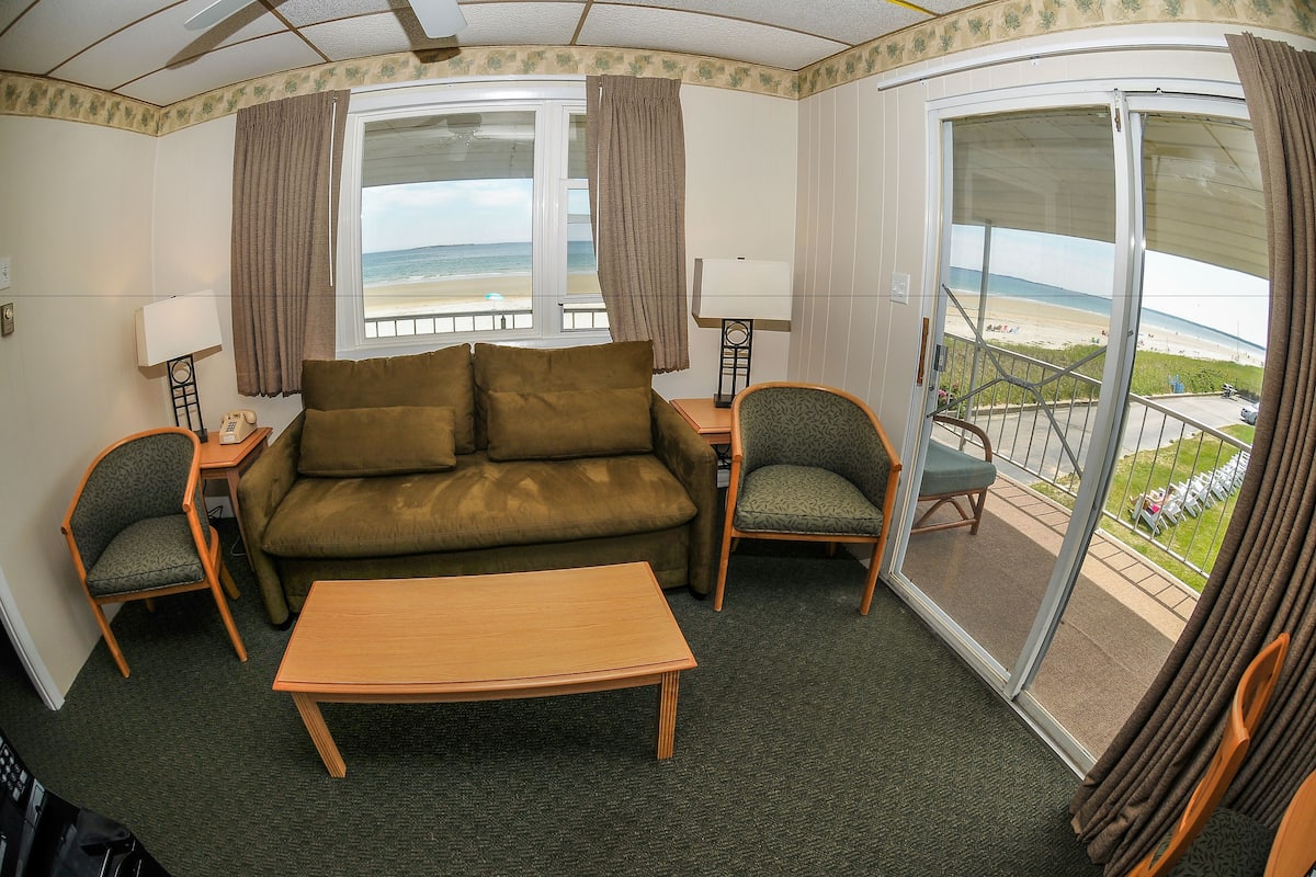 A room with a sofa, two chairs, a table, and a balcony with a view of the beach.