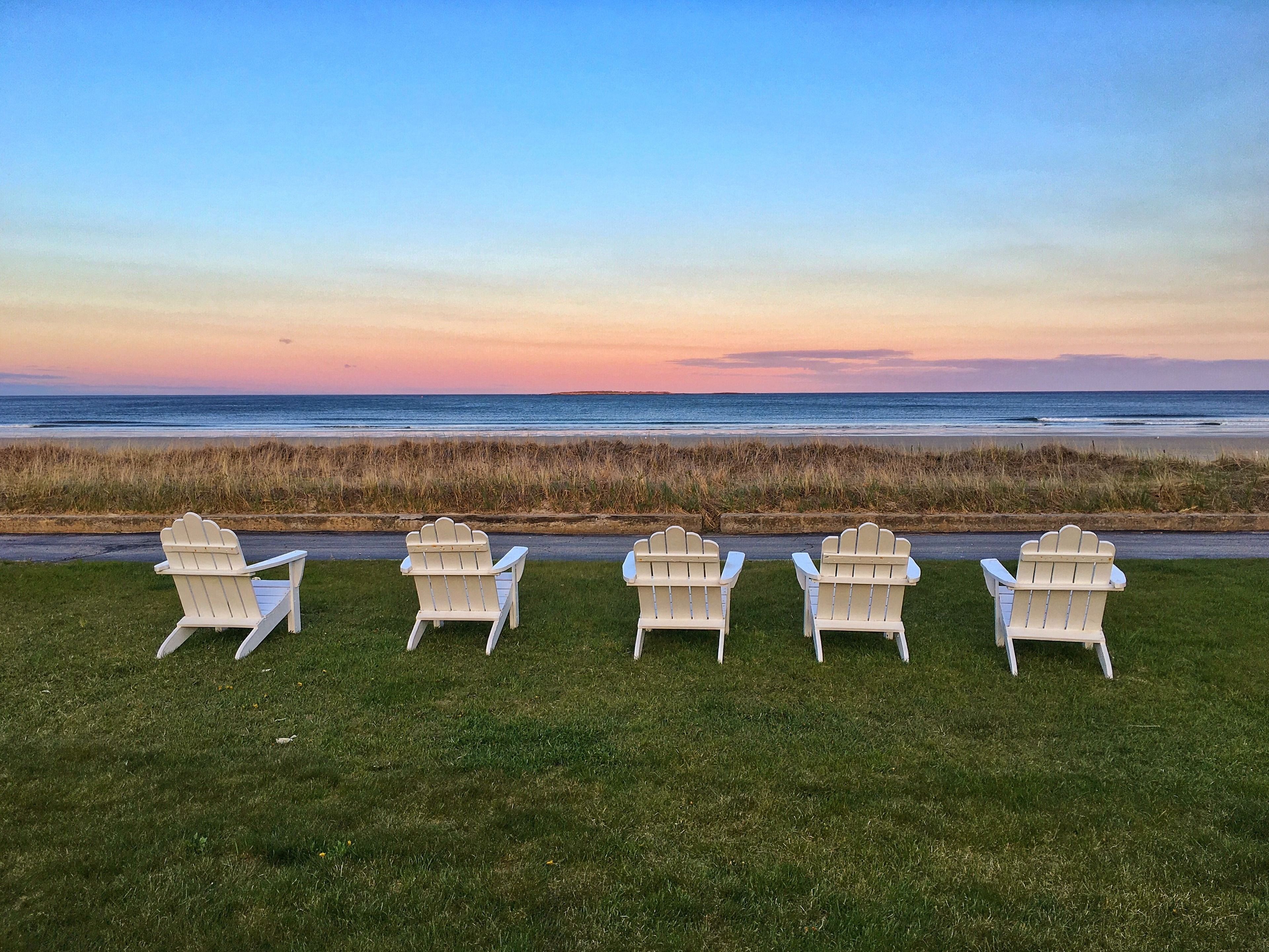 Quatre chaises Adirondack blanches sont alignées sur une pelouse, offrant une vue sur l’océan et la plage au coucher du soleil.