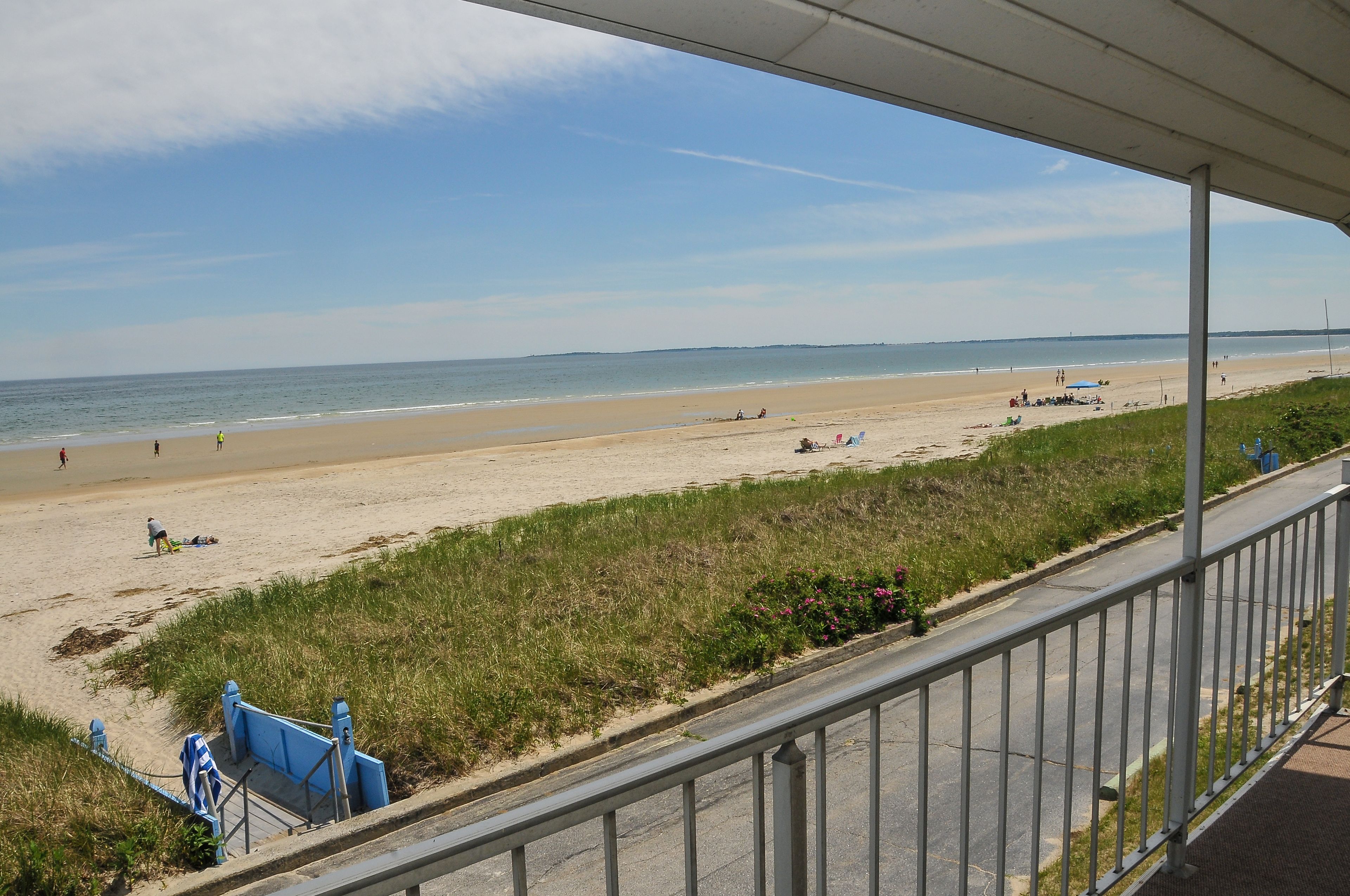 A beach view from a covered area with a railing, a grassy area, and a clear sky.