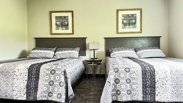 A hotel room with two beds, each with a patterned bedspread, a small bedside table with a lamp, and two framed pictures on the wall. - Colonial Valley Motel (Farmington)