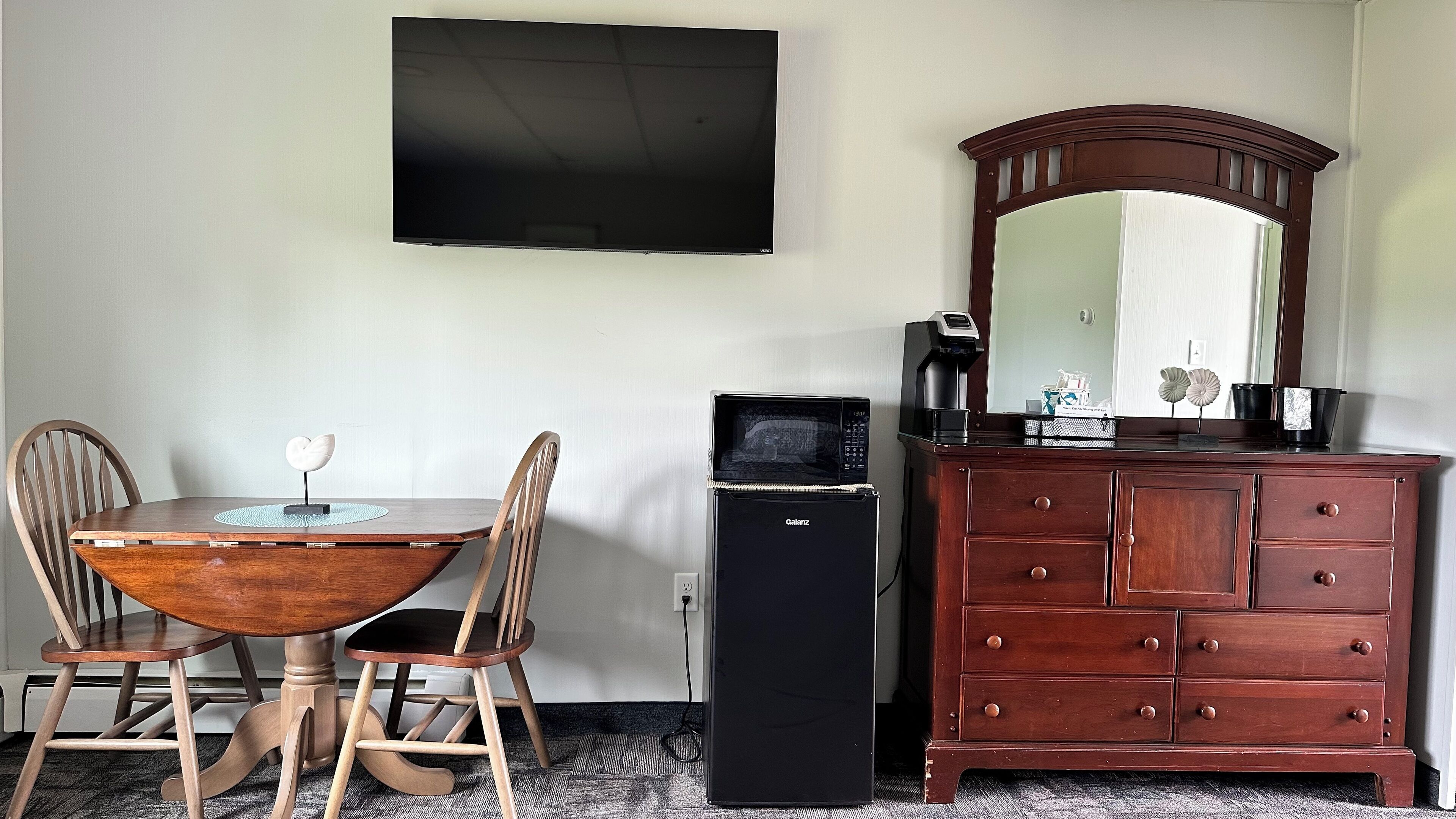 A room with a wooden dining table and chairs, a flat-screen TV mounted on the wall, a dark-colored refrigerator, and a wooden dresser with a mirror.
