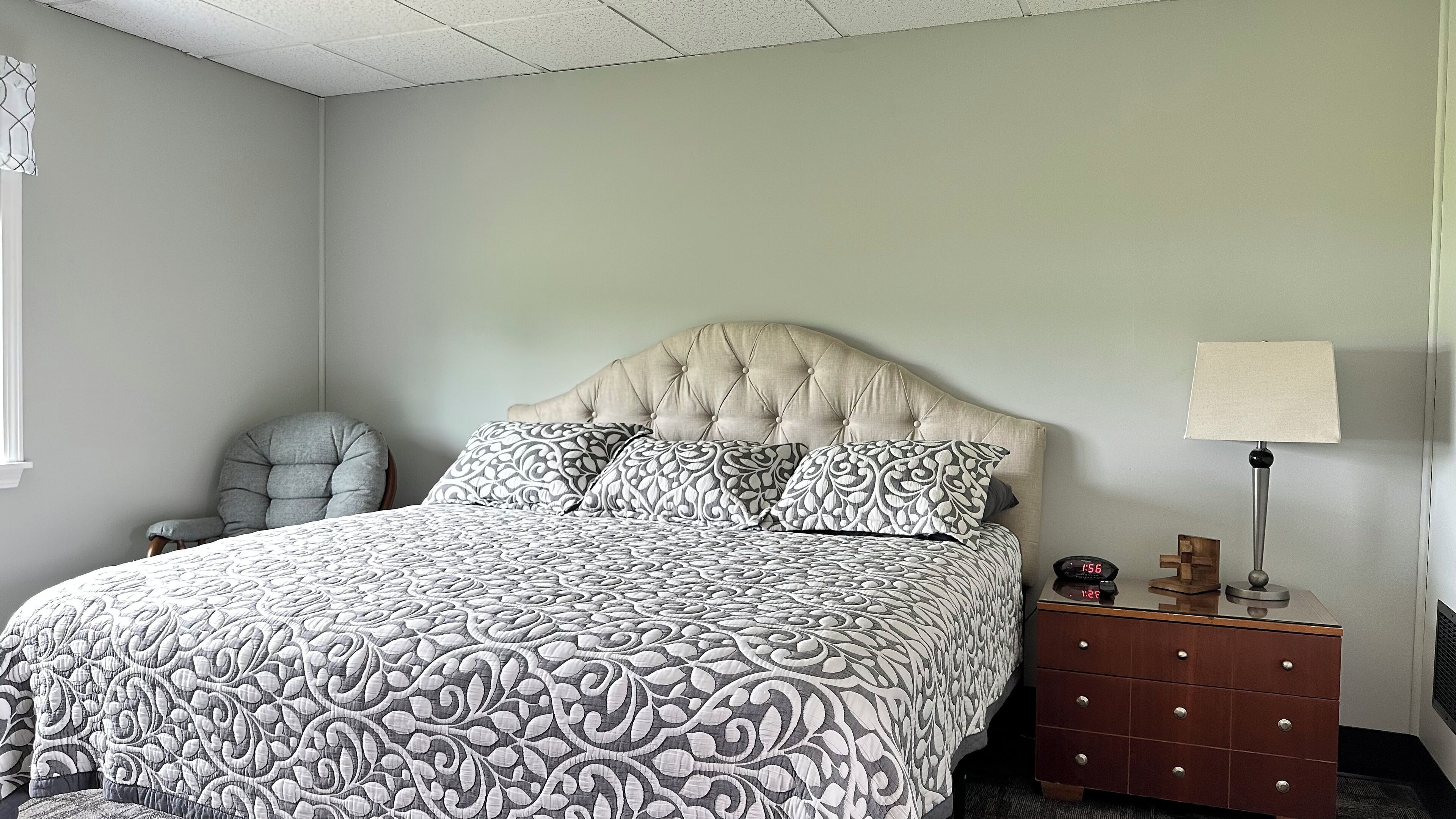 A bedroom with a tufted headboard, patterned bedspread, a bedside table with a lamp, and a chair.