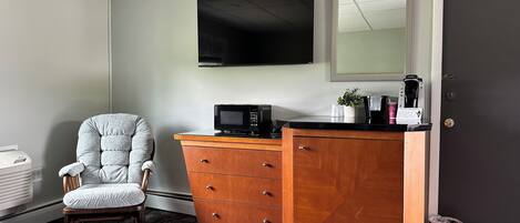 Habitación de hotel con un sillón gris, un aparador de madera con microondas, cafetera y una pequeña planta en maceta.